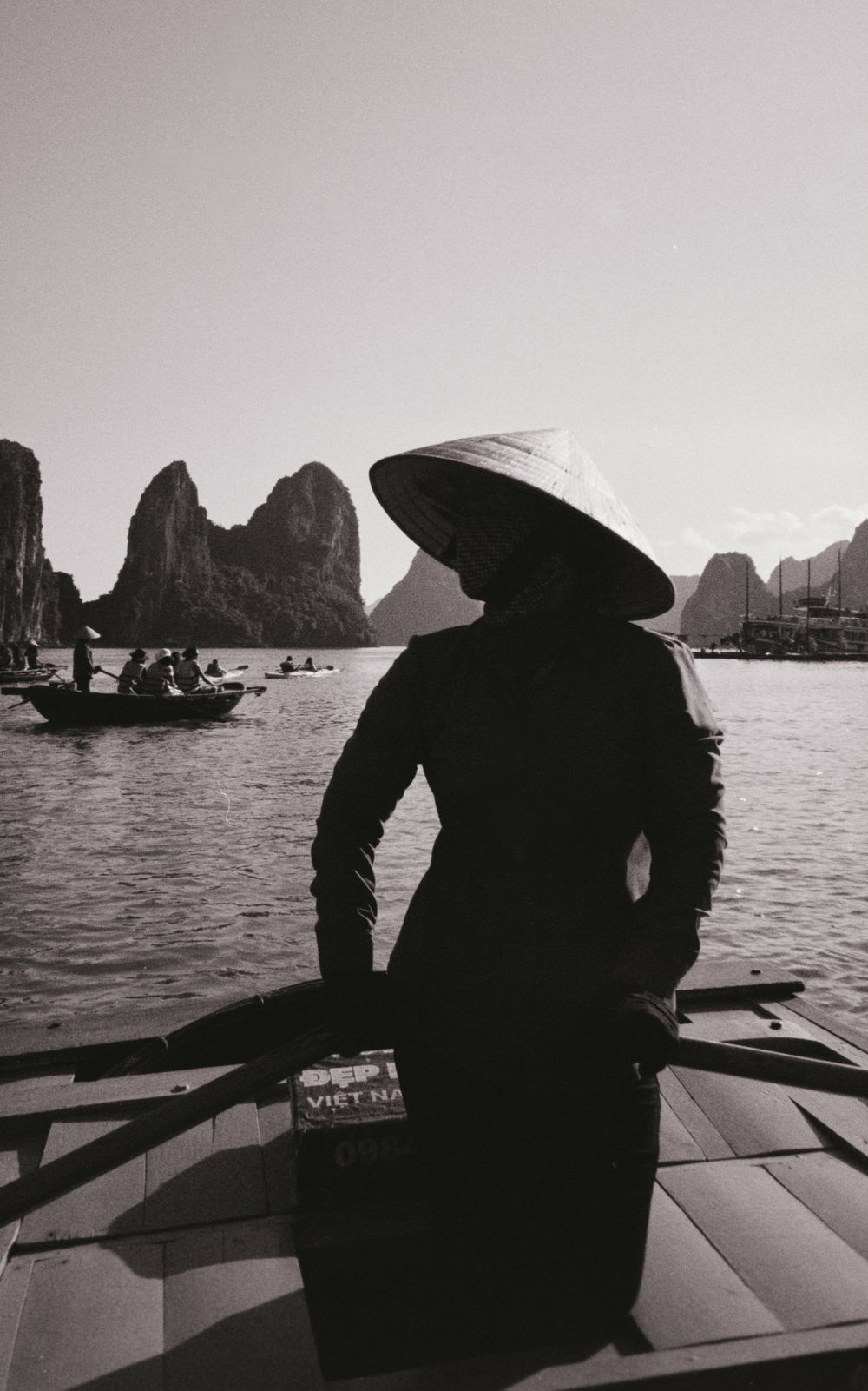A dramatic black and white shot of a silhouette in traditional Vietnamese hat aboard a boat surrounded by karst formations.
