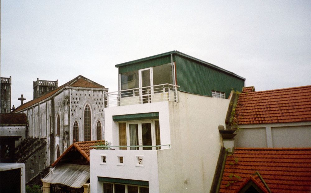 A quirky white and green two-story building with slanted architecture stands among traditional red-tiled roofs.