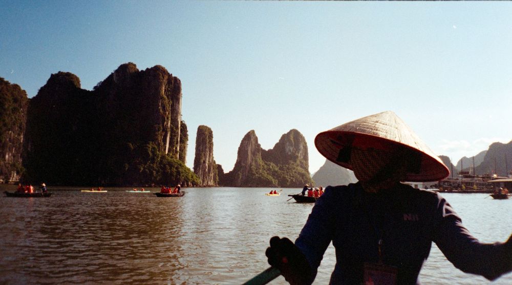 A silhouetted figure wearing a conical hat rides a boat through dramatic limestone karsts in Halong Bay, Vietnam.