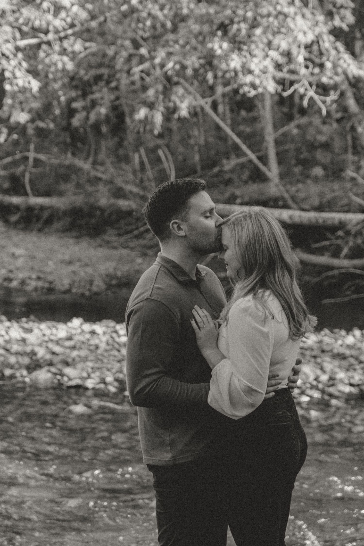Romantic black and white photograph of a couple embracing by a stream in nature.