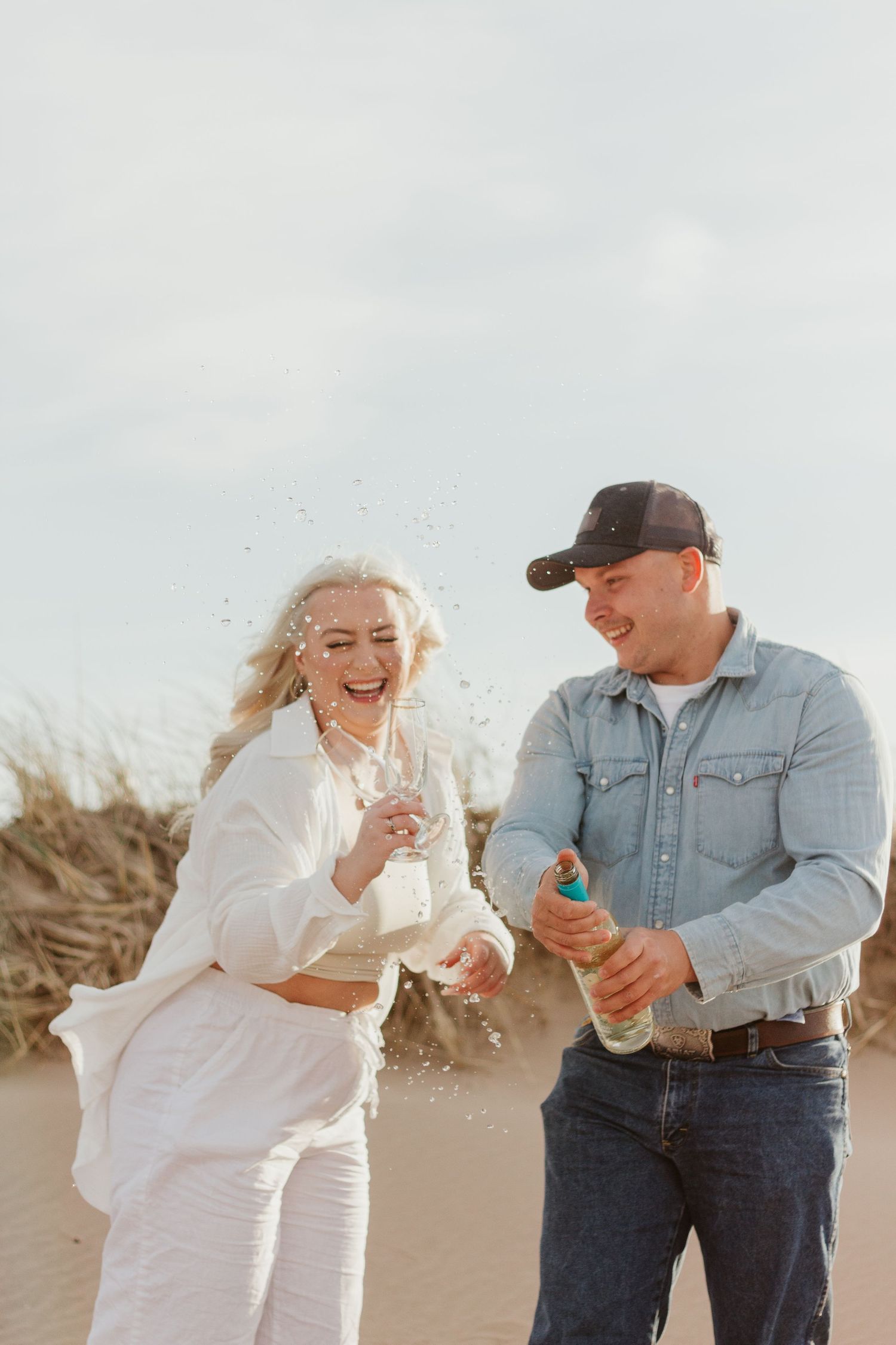 A playful couple pops champagne together on a beach at sunset.