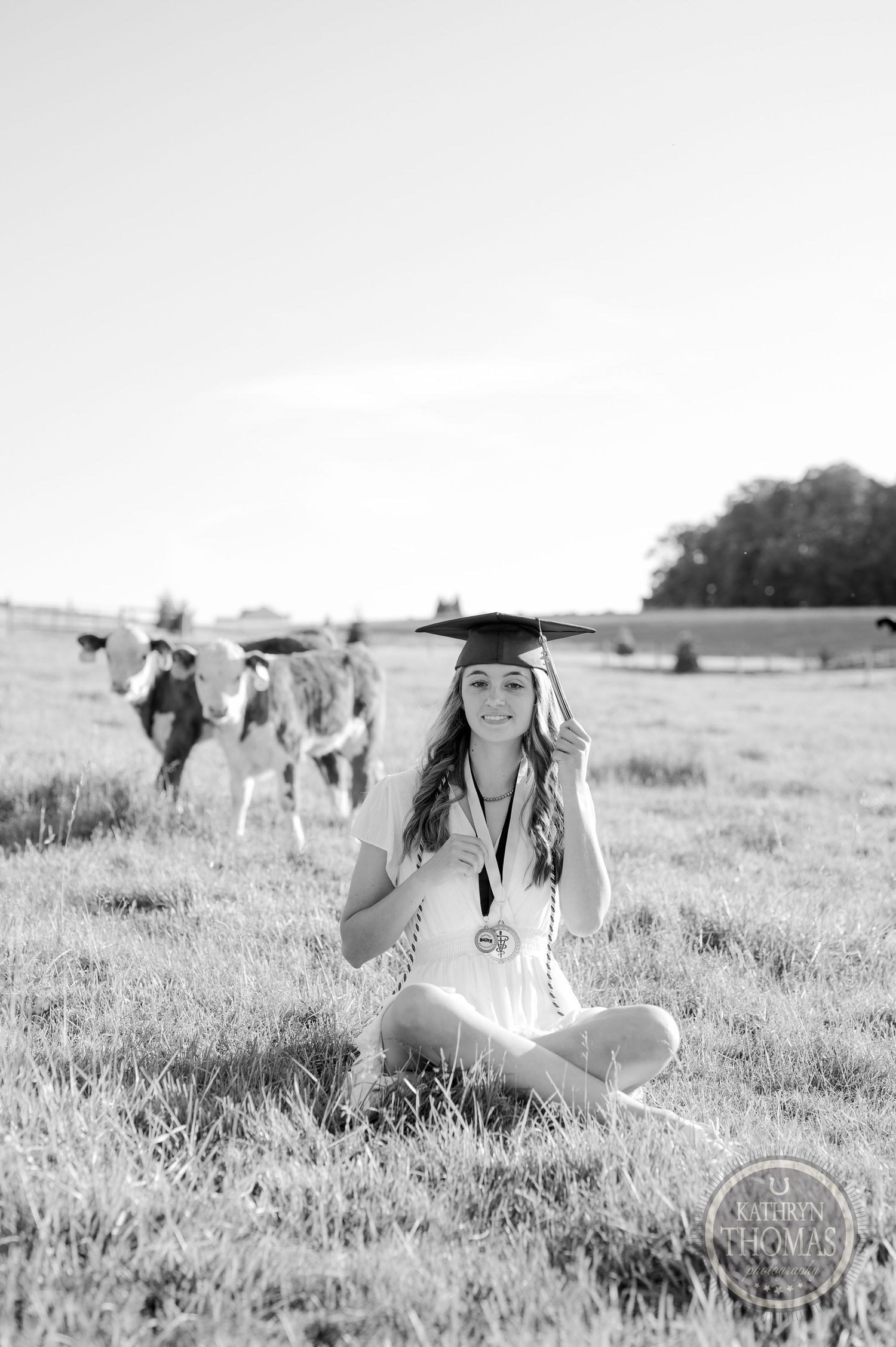 Black and white portrait of graduate sitting in field with cattle in background.