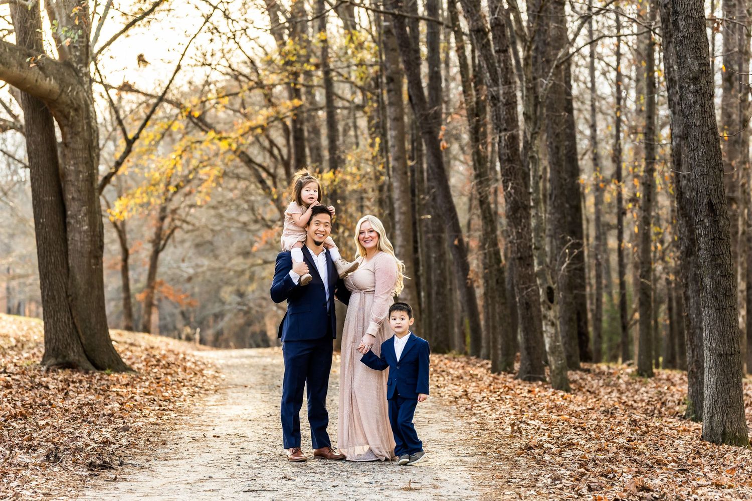 Family portrait on tree-lined autumn path with falling leaves.