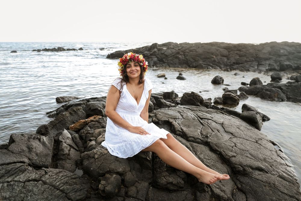 Woman in white dress poses on black volcanic rocks near ocean.