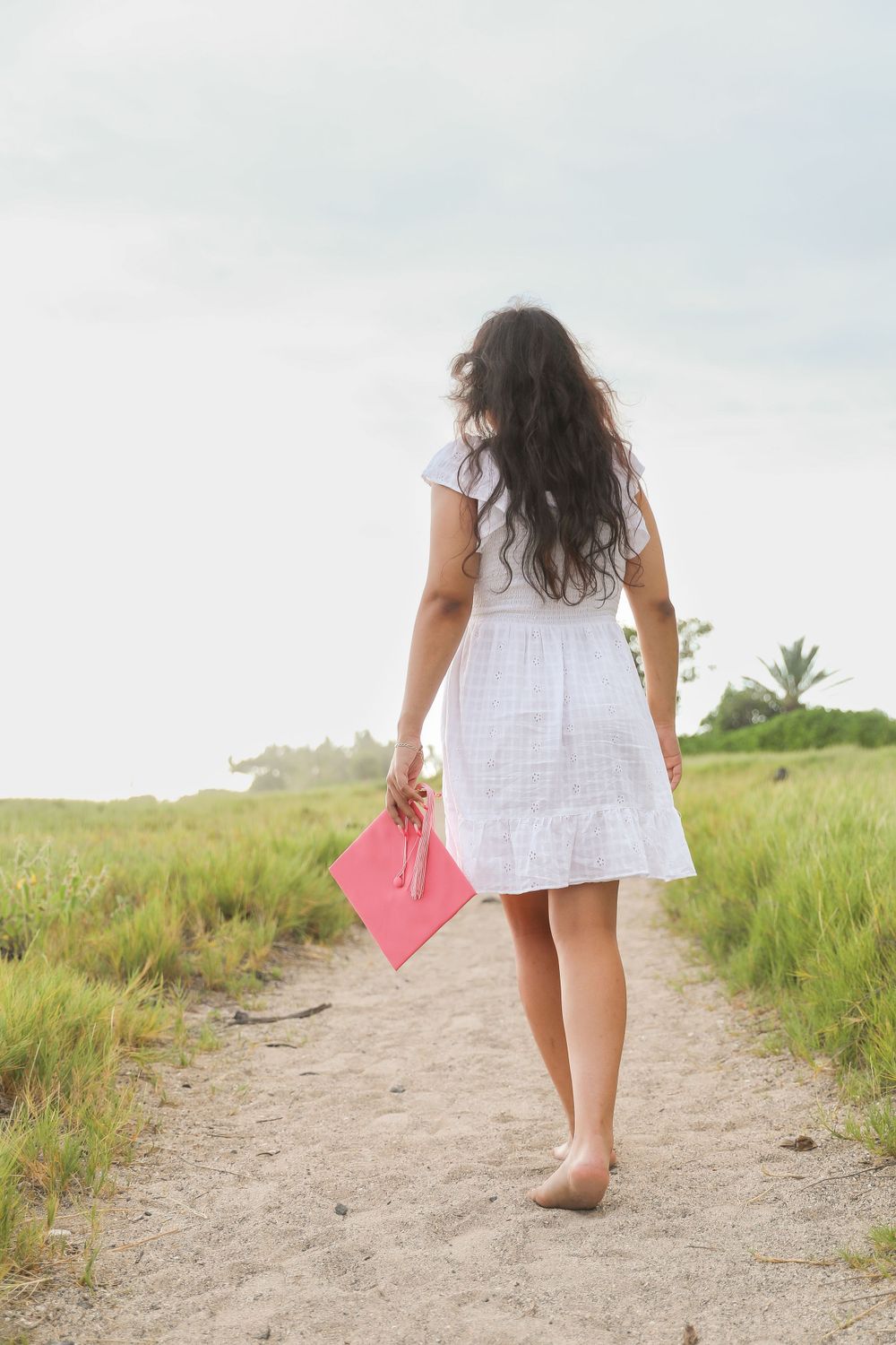 Woman in white dress walks barefoot on dirt path carrying pink graduation cap.