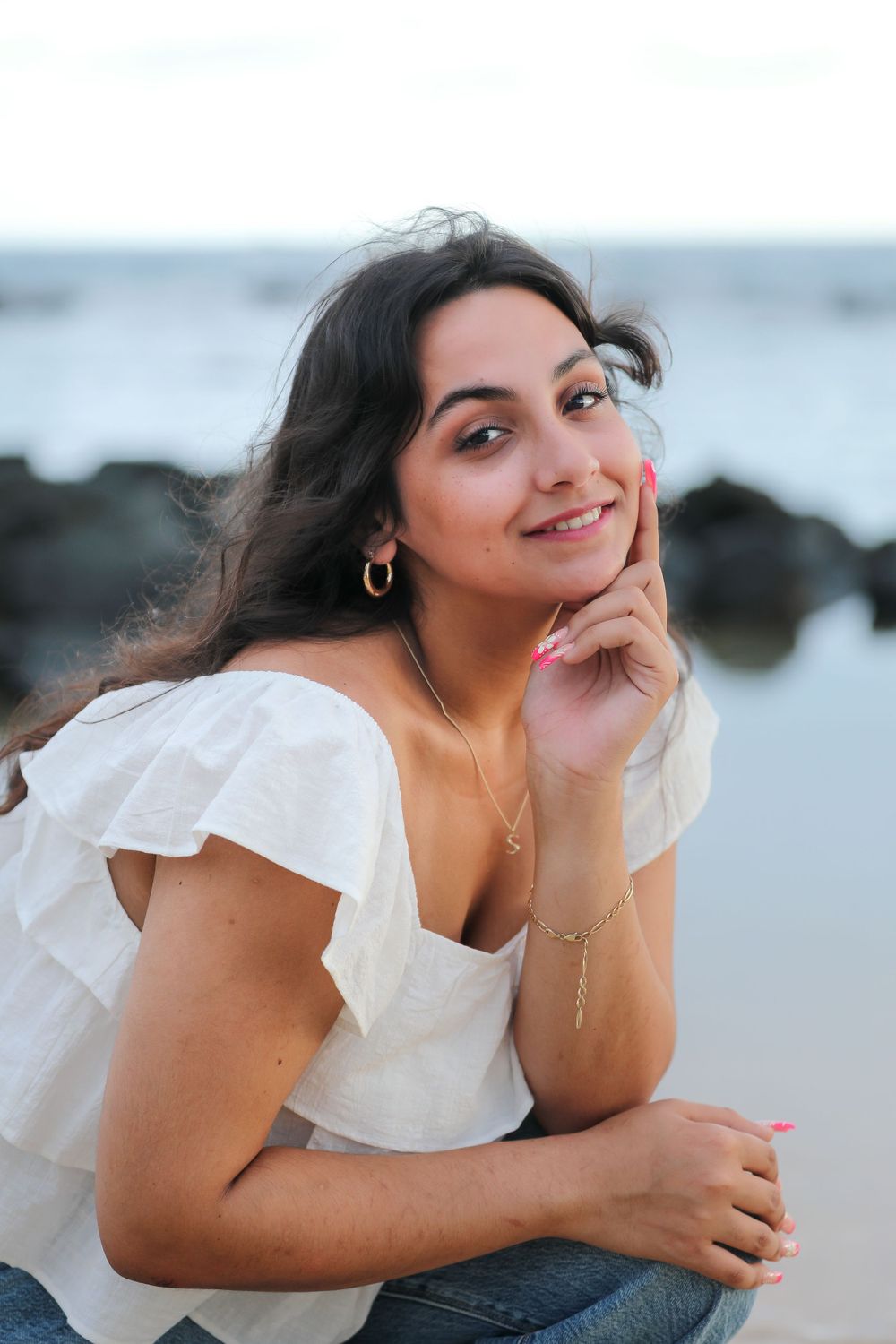 Casual seaside portrait with white ruffled blouse and delicate jewelry against ocean rocks.