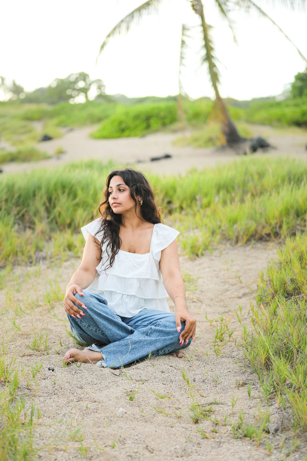 Woman in ruffled white top and jeans sits on sandy path with palm trees.