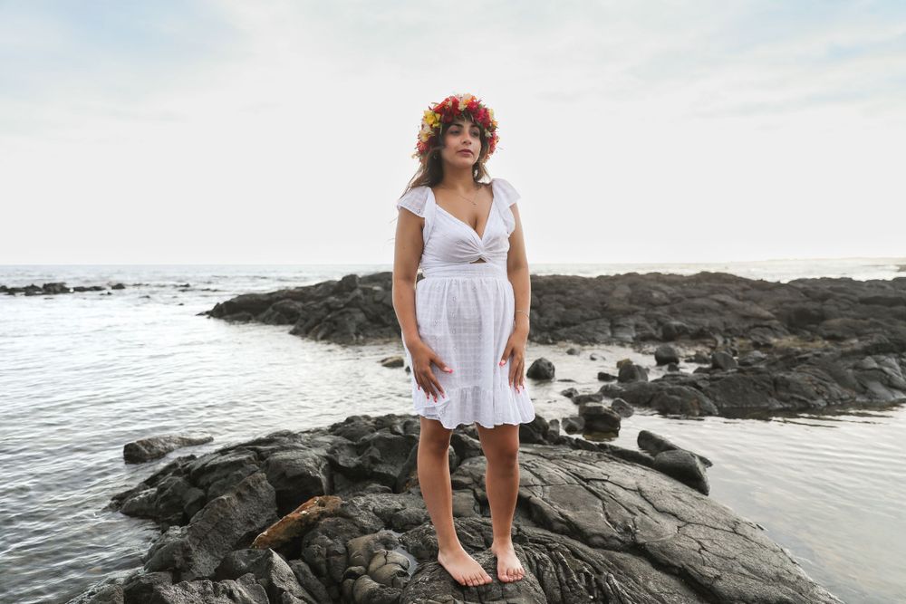 Woman in white dress stands on volcanic rocks by ocean.