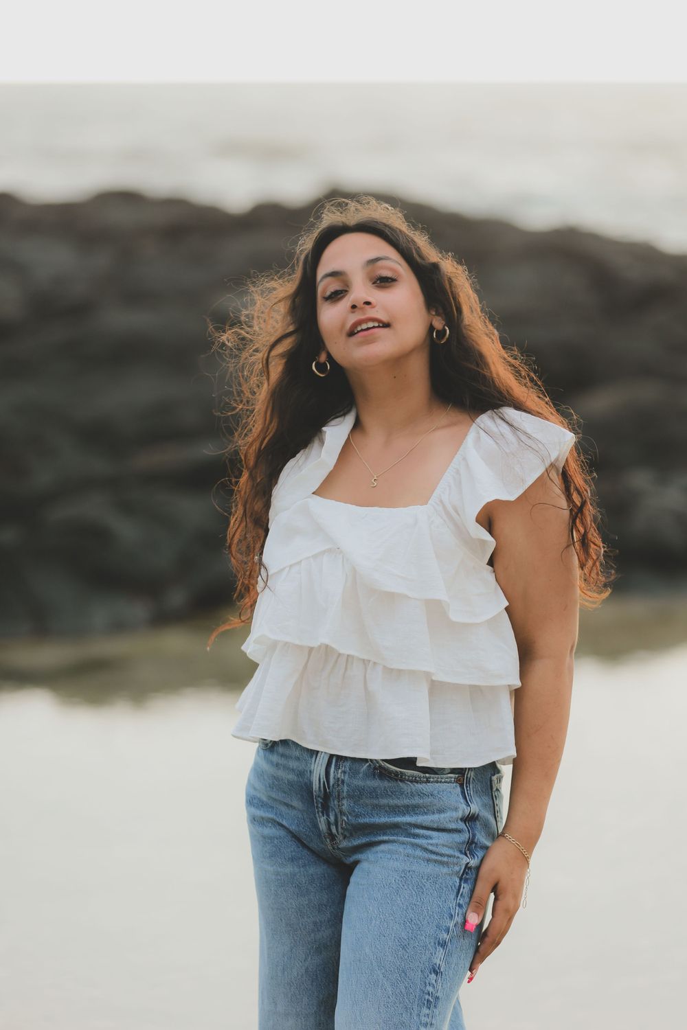 A casual beach portrait featuring a white ruffled top and blue jeans against a rocky coastal backdrop.