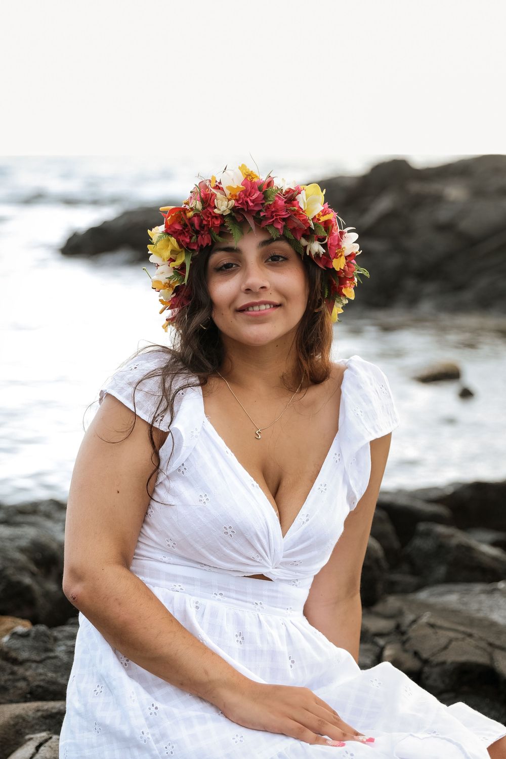 Woman in white dress wears colorful flower crown while sitting on coastal rocks.