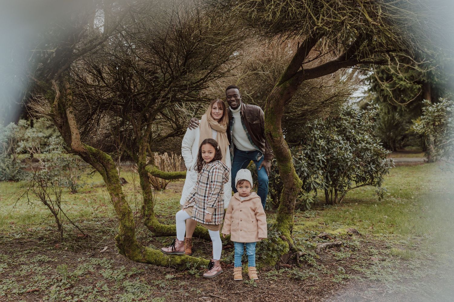 Family of four poses together under twisted tree branches in misty park.