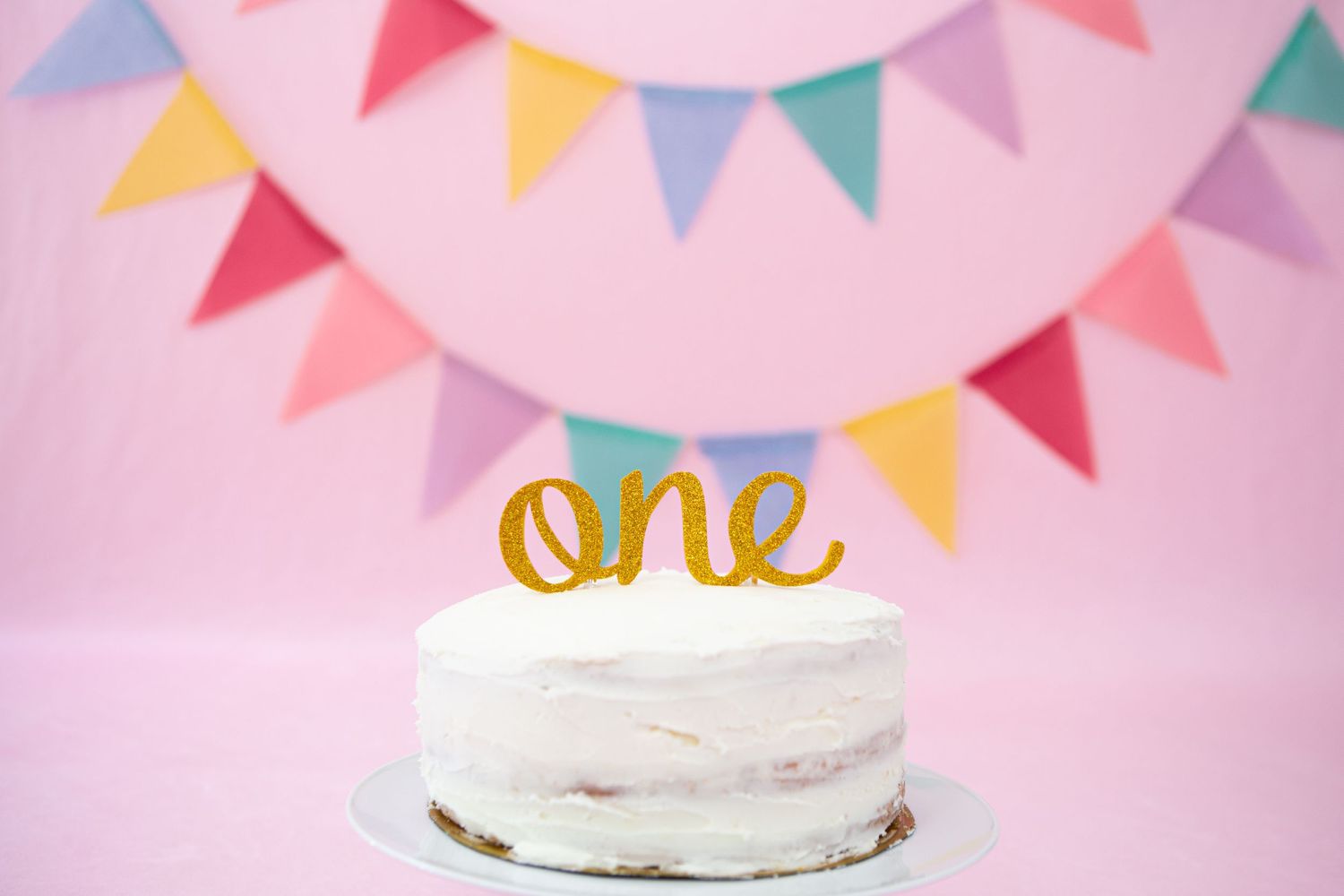 White birthday cake with gold 'one' topper and colorful triangle banner backdrop.