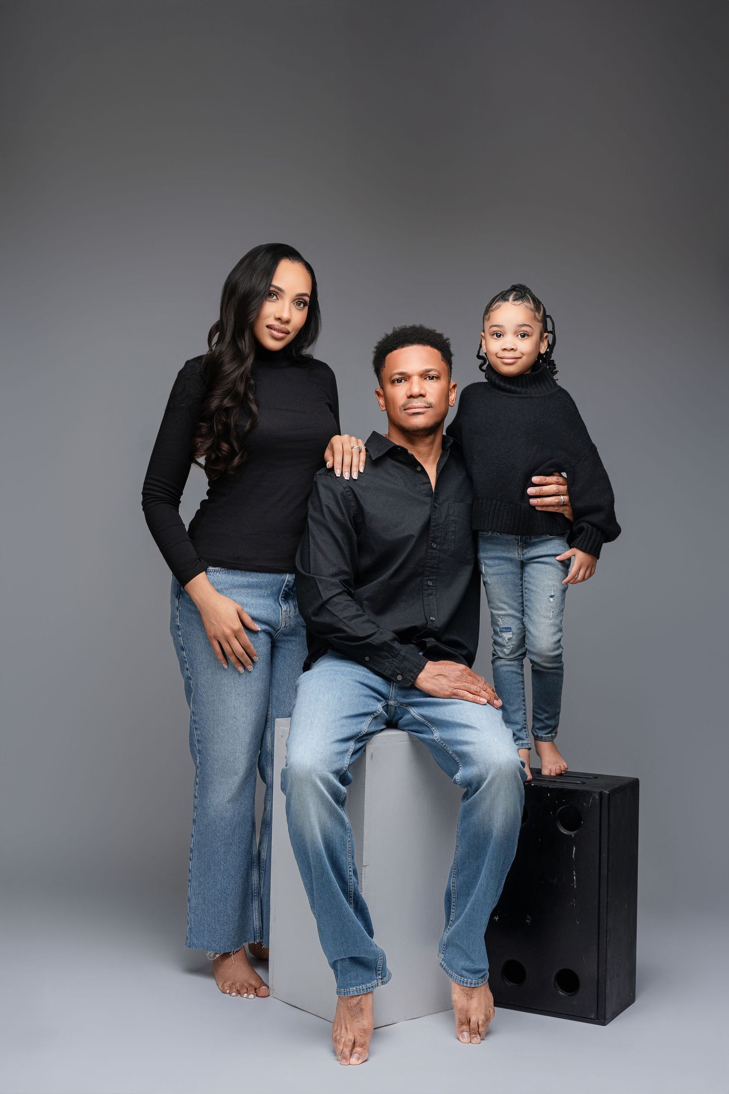 Family wearing black tops and blue jeans poses casually together on black cube in studio setting against gray backdrop.
