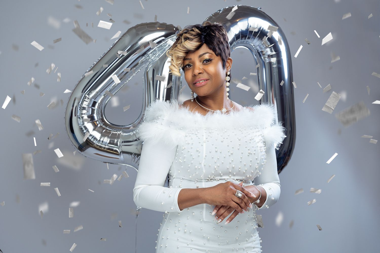 A bride in a white lace dress poses in front of silver balloon decorations while confetti falls around her.
