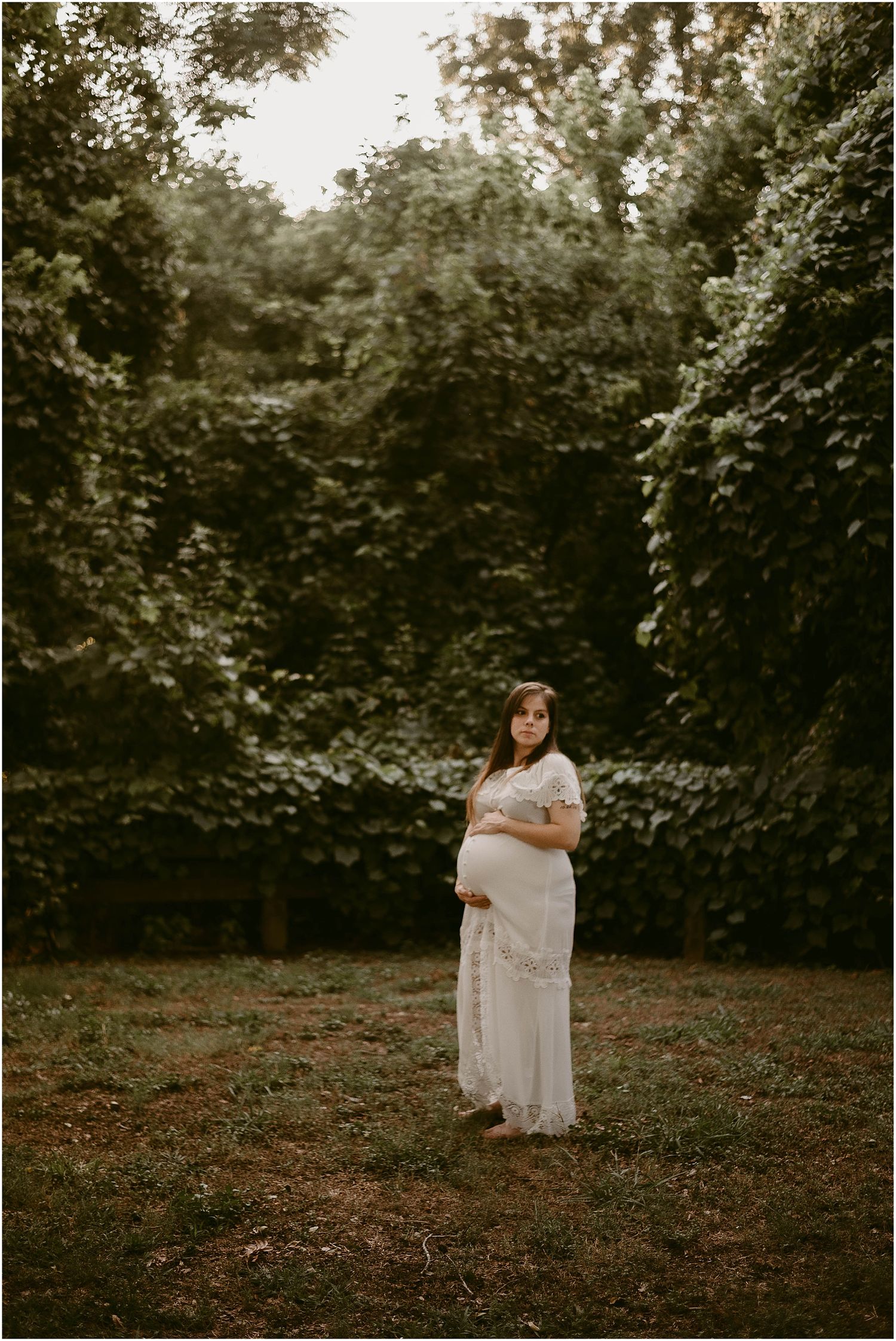 Maternity photo of pregnant woman in white lace dress standing in garden with greenery backdrop.