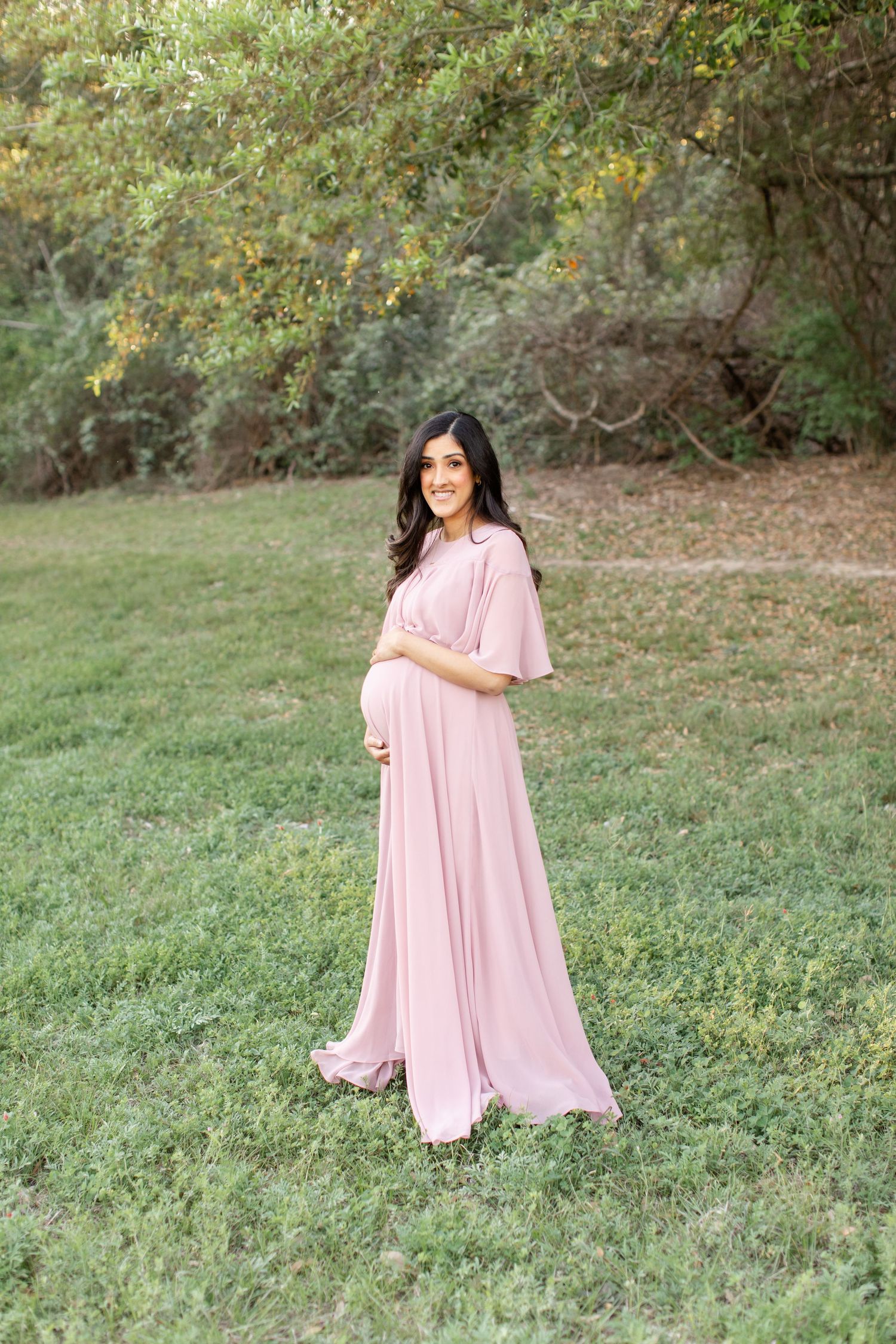 A flowing pink maternity dress is worn gracefully in an outdoor garden setting.