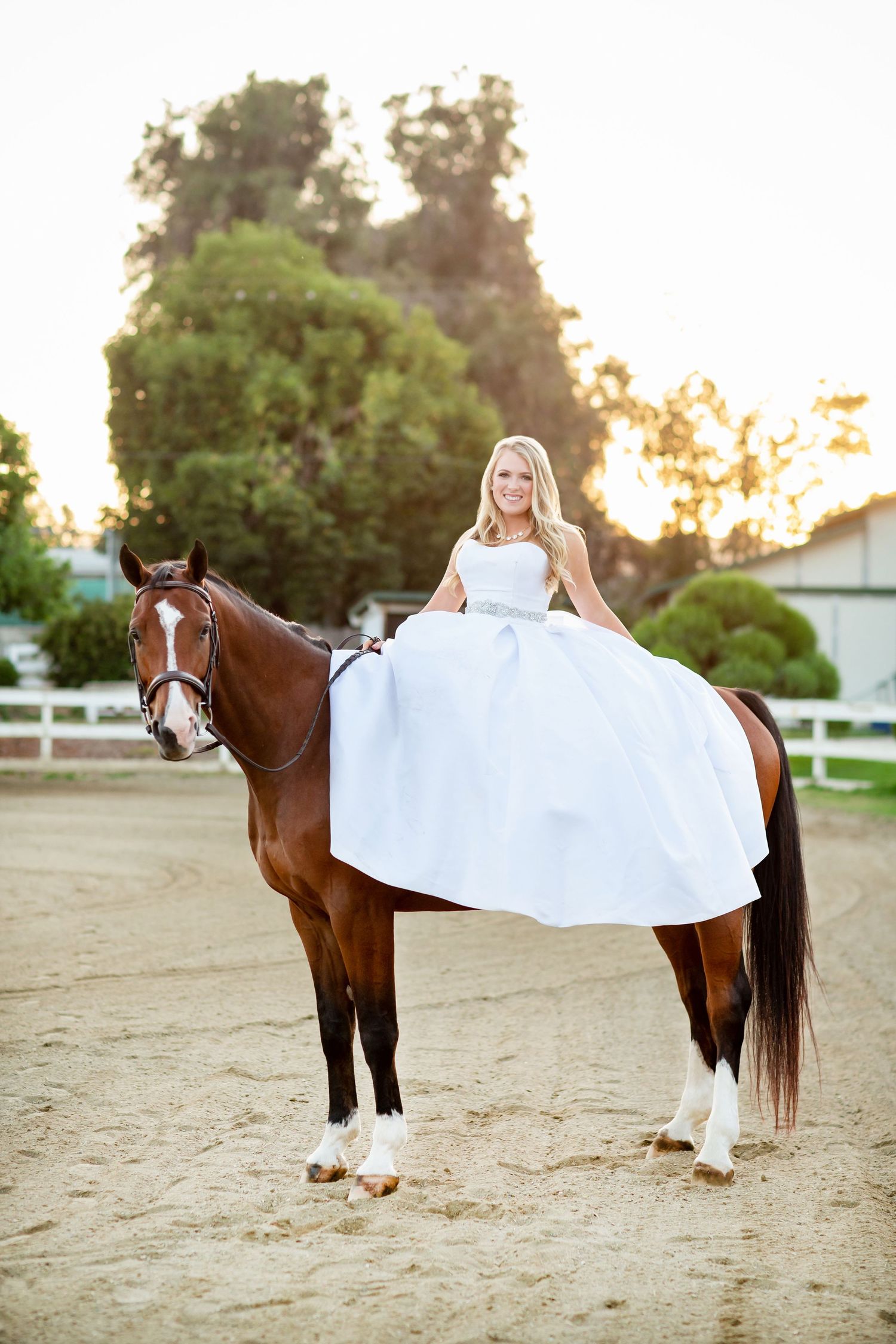 A person in a white formal dress sits atop a brown horse at sunset on a ranch.
