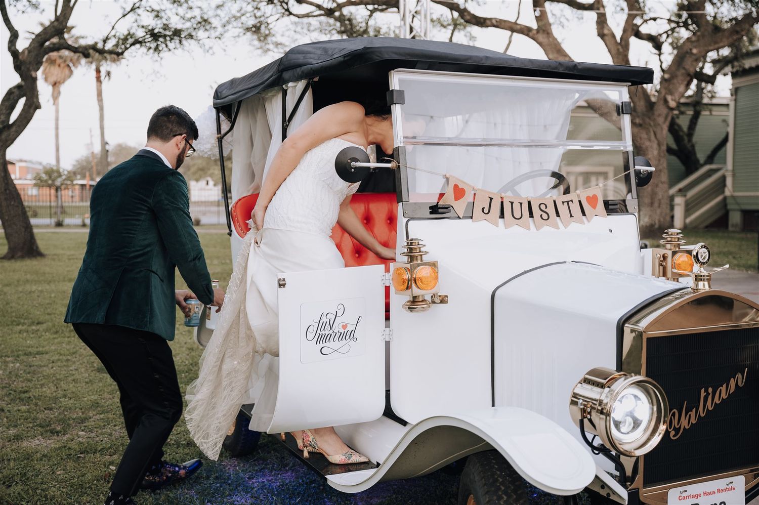 A vintage white wedding car with couple entering on a grassy field.