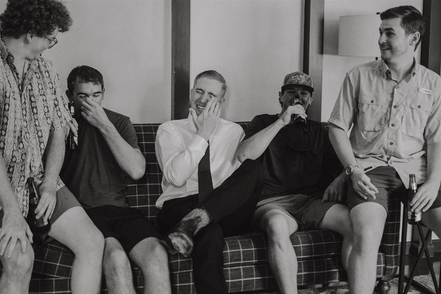 Groomsmen share a lighthearted moment on a plaid couch in black and white photography.
