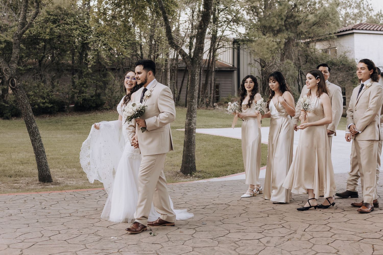 Wedding party in beige attire watches couple embrace during outdoor celebration.