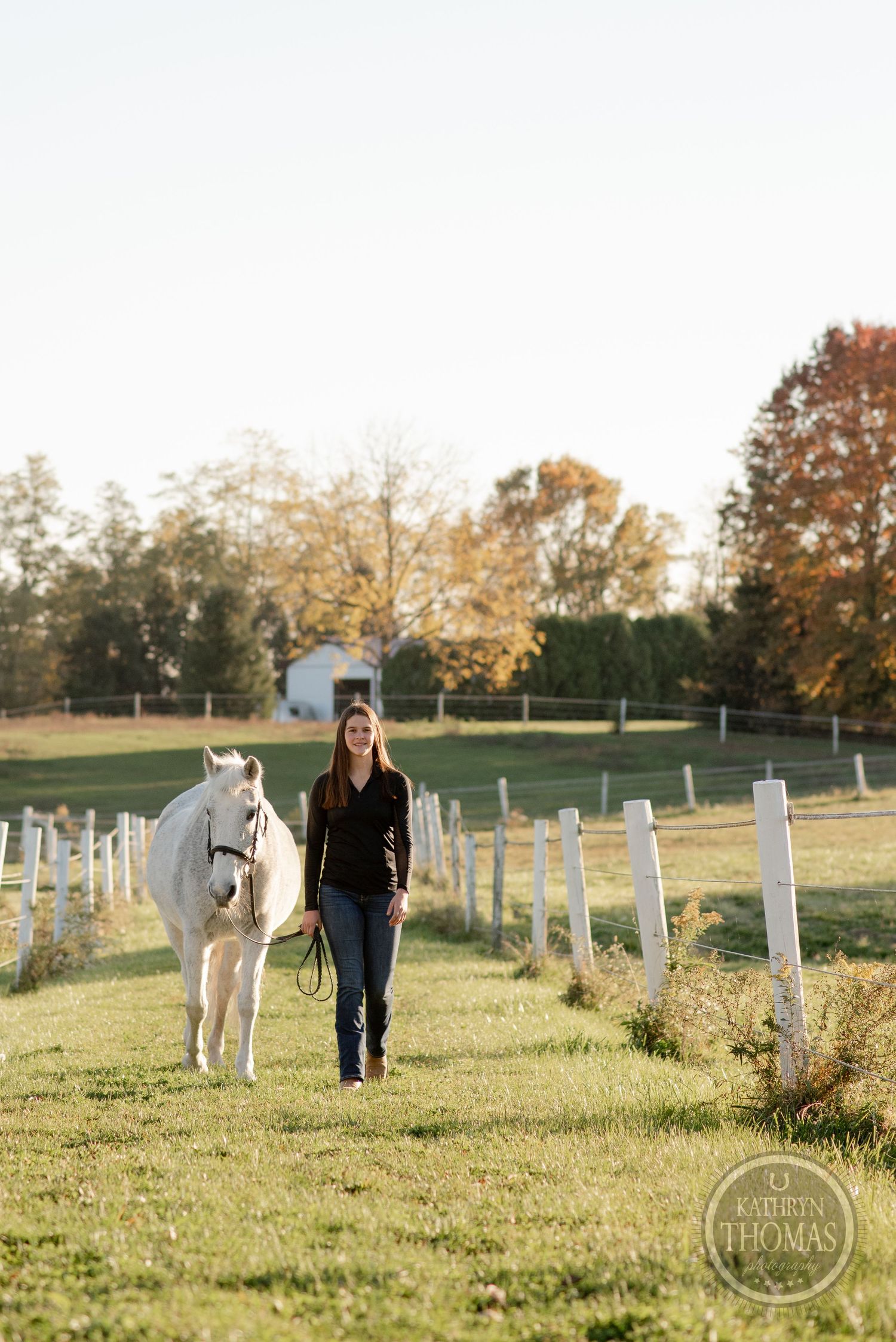 Person walking with white horse along fence line during golden hour.