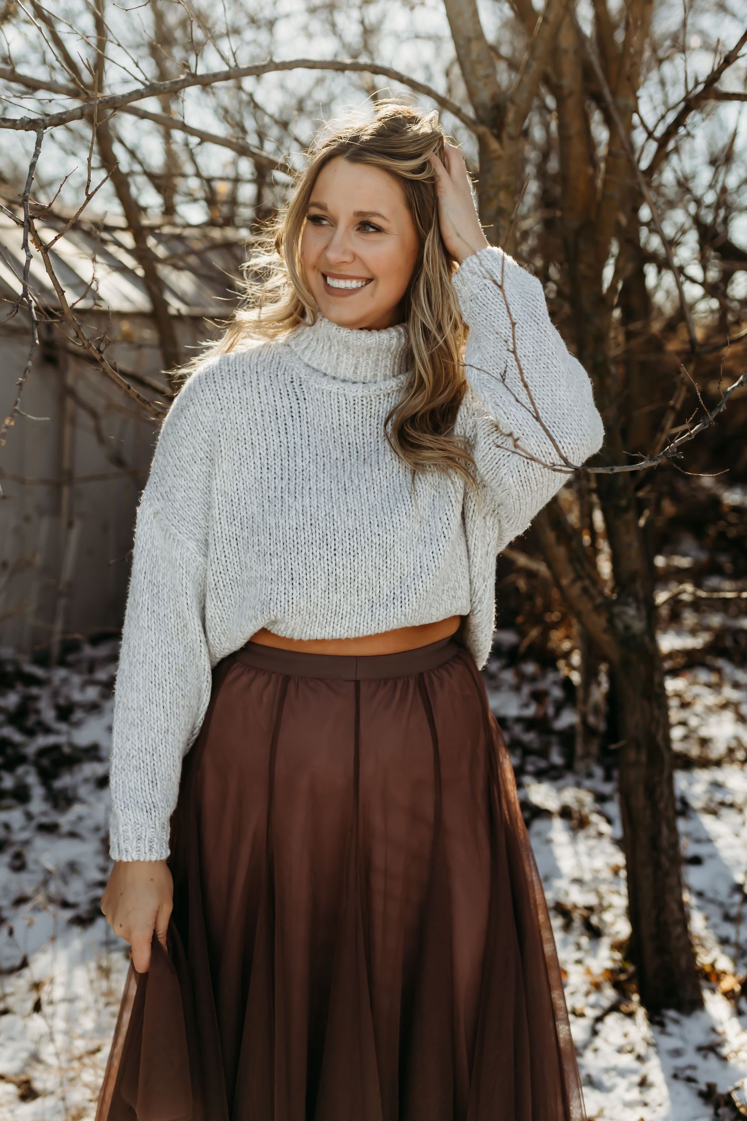 Woman in white sweater and brown skirt standing in snowy winter scene.