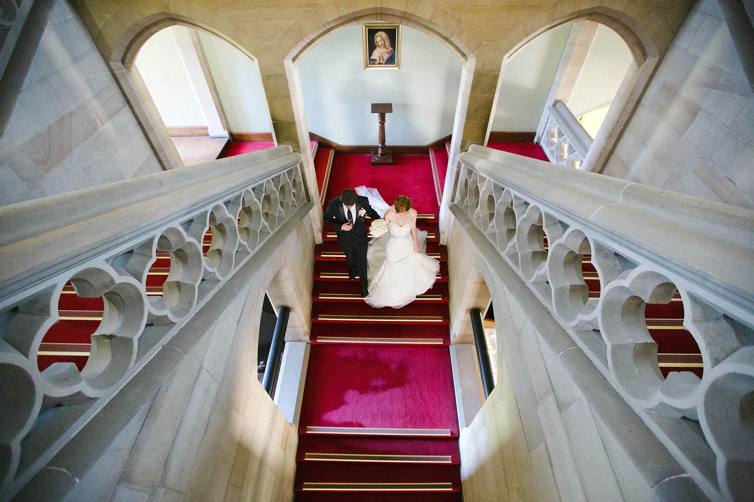 Wedding dress details flowing down elegant white marble stairs with red carpet.