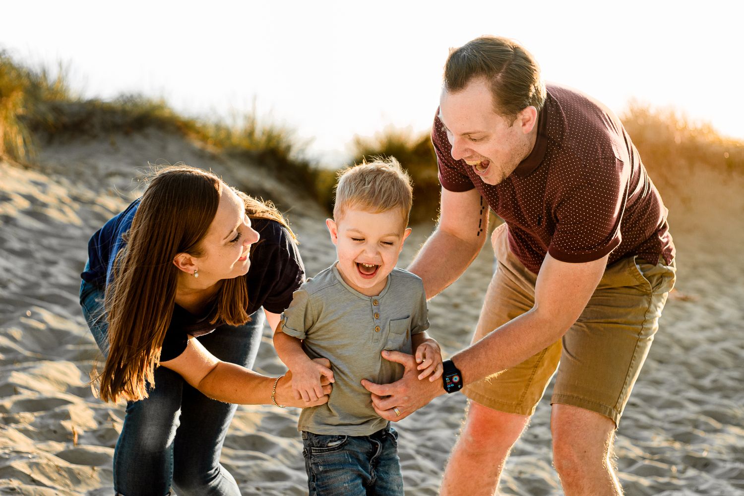 On a beach on Hayling Island parents tickle their three year old in the sand.