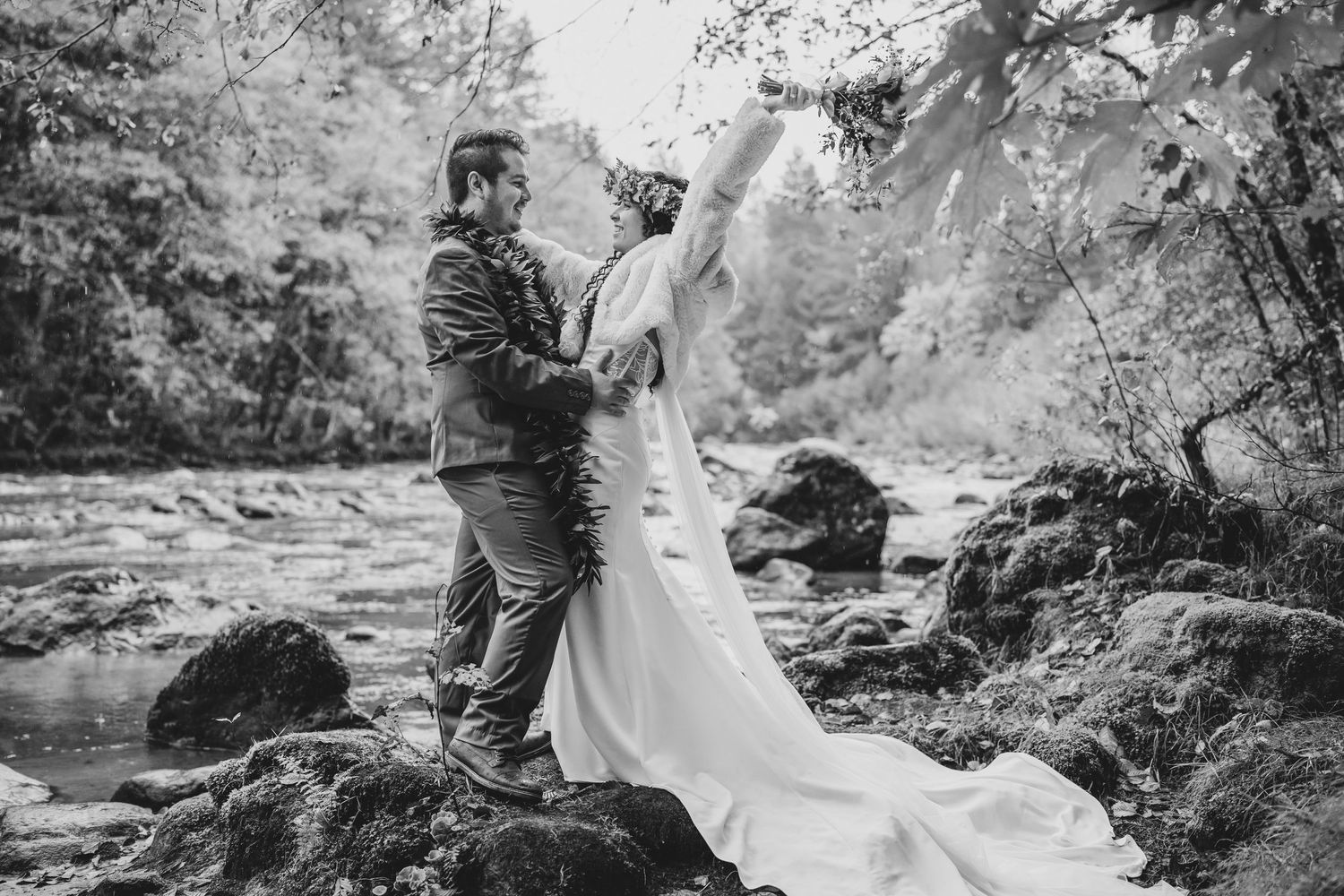 A couple stands on rocky terrain by a river in a romantic black and white wedding photo.