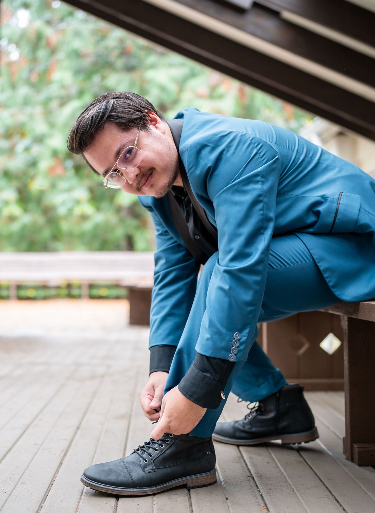 Someone in a blue suit adjusts their shoe while sitting on a wooden bench outdoors.