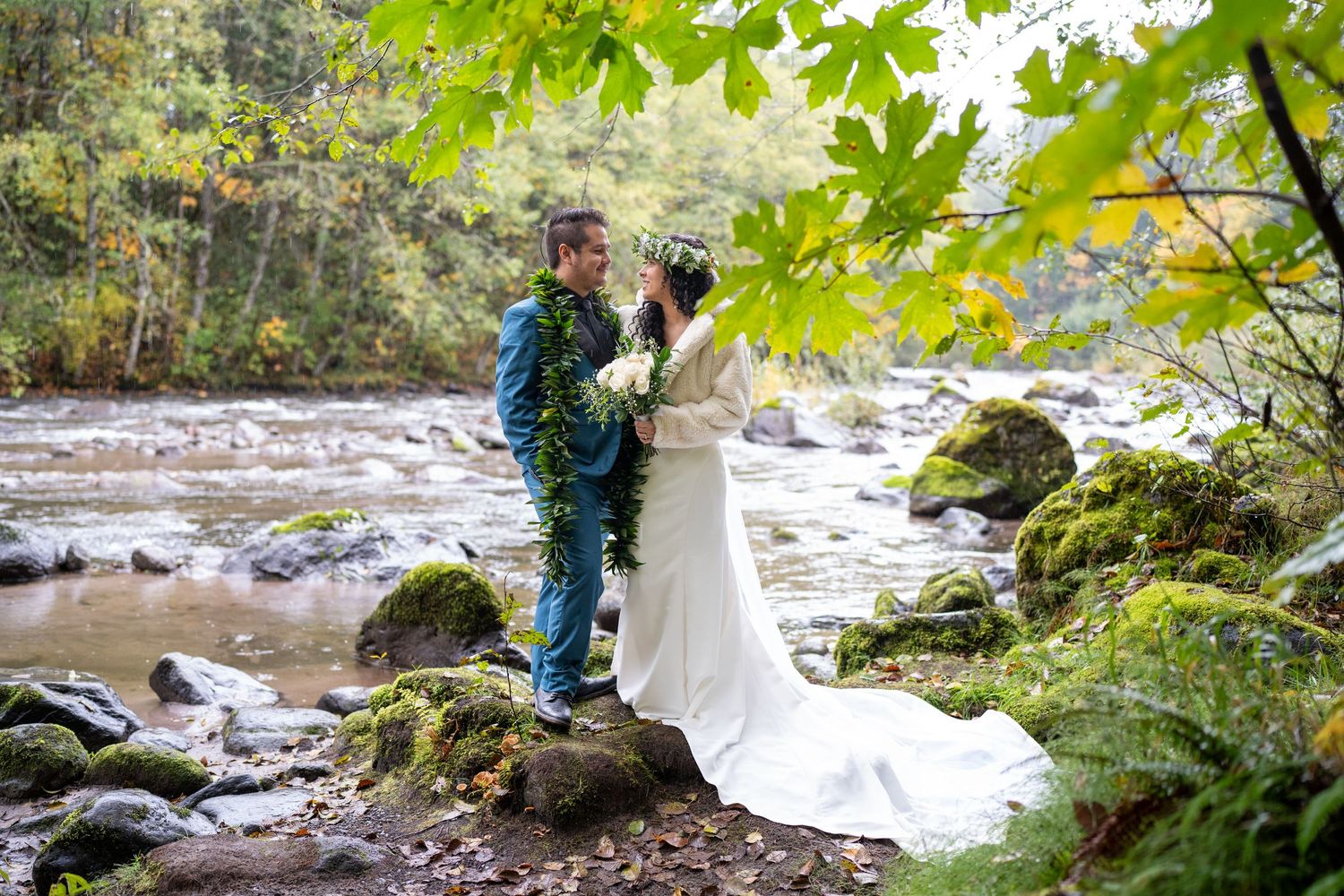 A couple stands on mossy rocks by a rushing river surrounded by lush greenery during a wedding photoshoot.