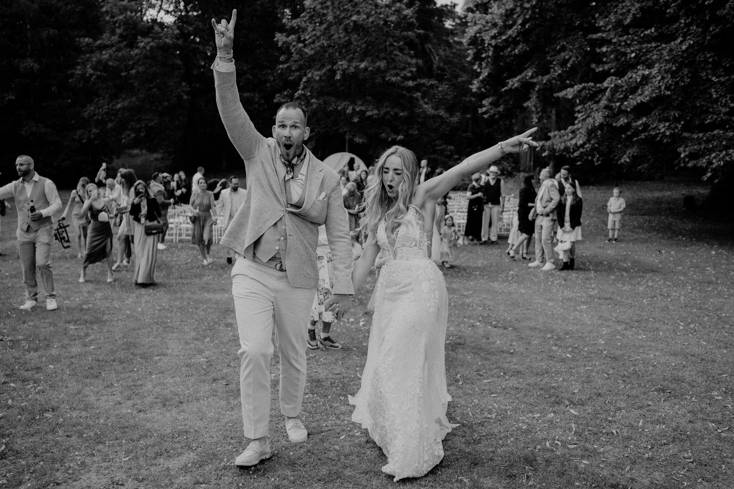 A joyful wedding couple celebrates on a grassy lawn while wedding guests look on in this black and white photograph.