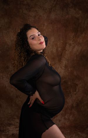 Dramatic maternity portrait in black sheer dress against brown textured backdrop.
