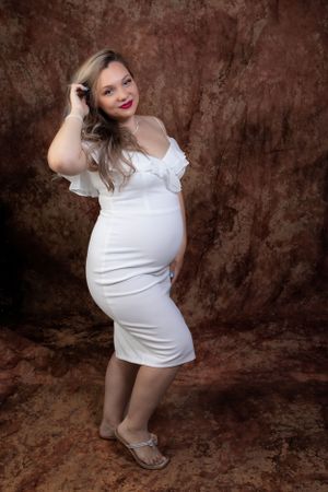 A maternity portrait against a dark brown backdrop featuring a white form-fitting dress.