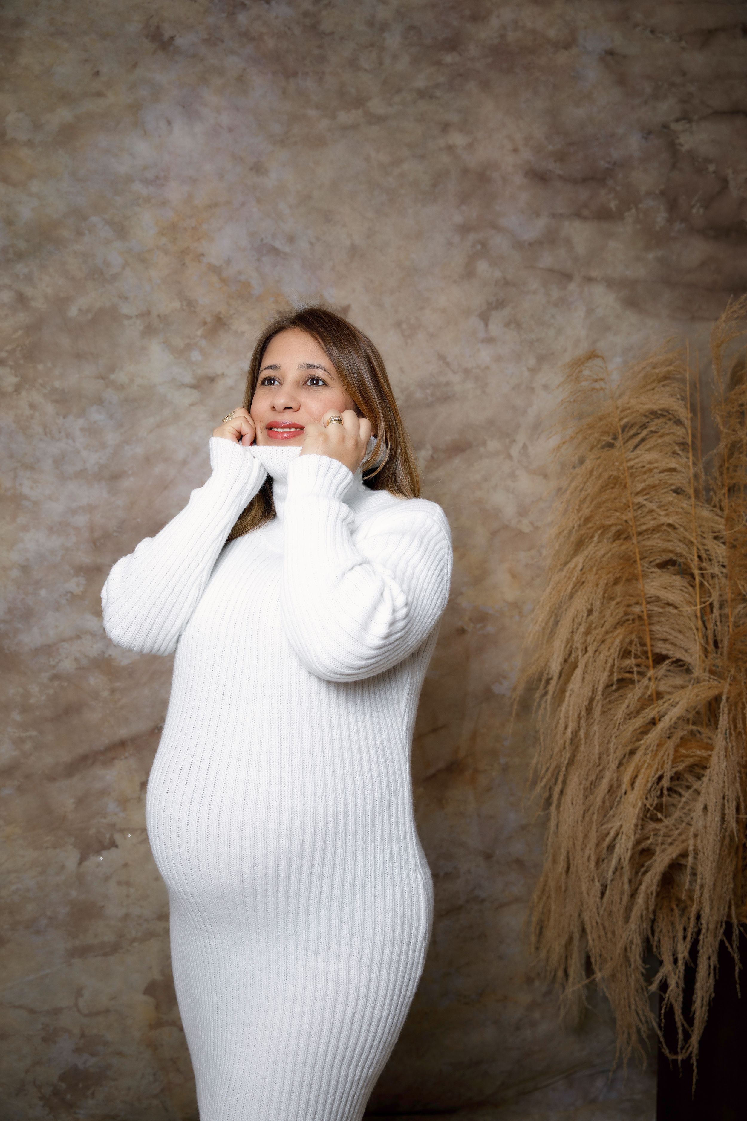 Maternity portrait of woman in white sweater dress posing next to pampas grass.