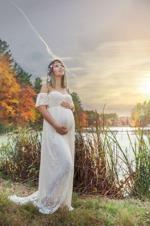 An expectant mother in white lace poses by a lake during autumn sunset.