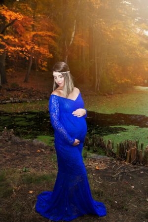 A maternity photo of a woman in a blue lace gown against fall foliage.