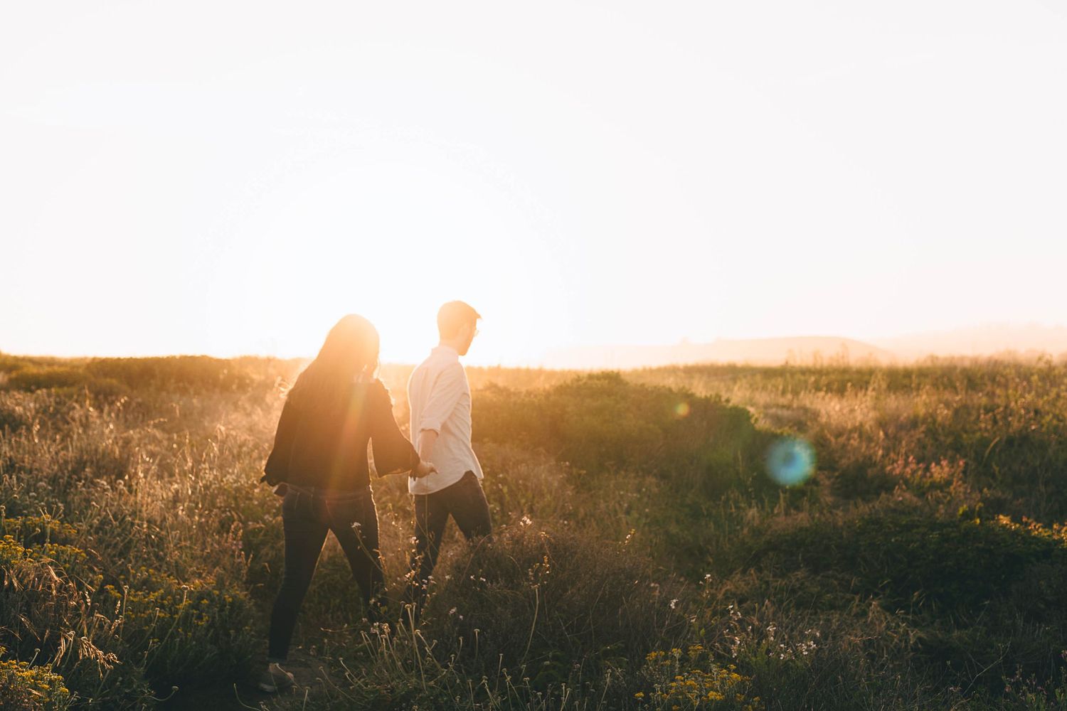 Two people walking hand in hand through a sunlit field at golden hour.