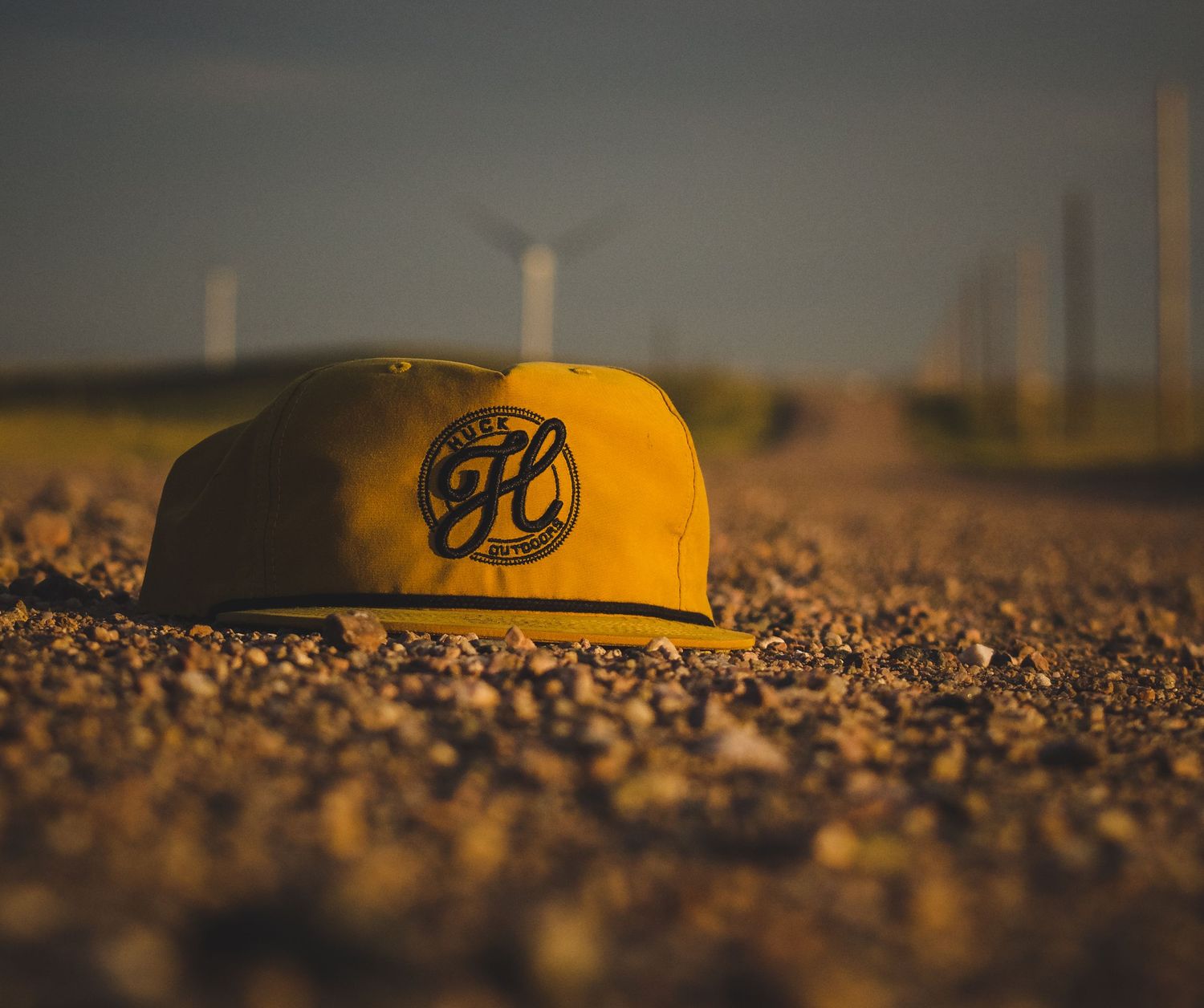 A yellow baseball cap with FC logo lies on gravelly ground at sunset.