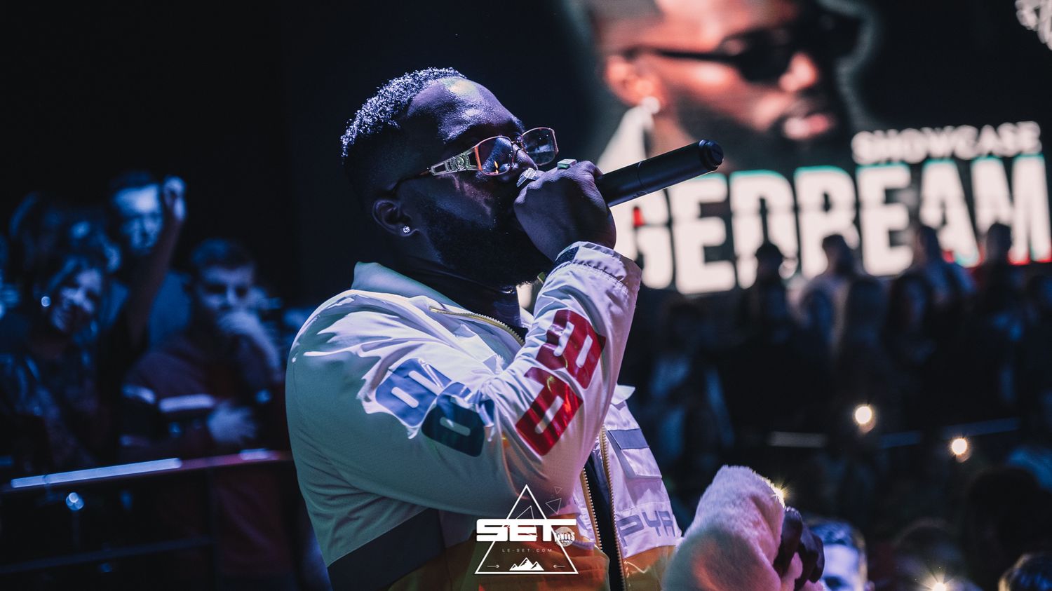 Rapper performing on stage with microphone at a live concert event with crowd in background.