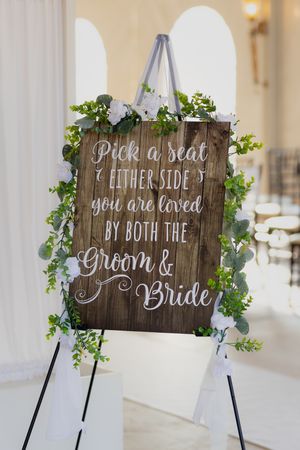 Rustic wooden wedding sign decorated with greenery reads 'Pick a seat, you are loved by both the bride & groom'.