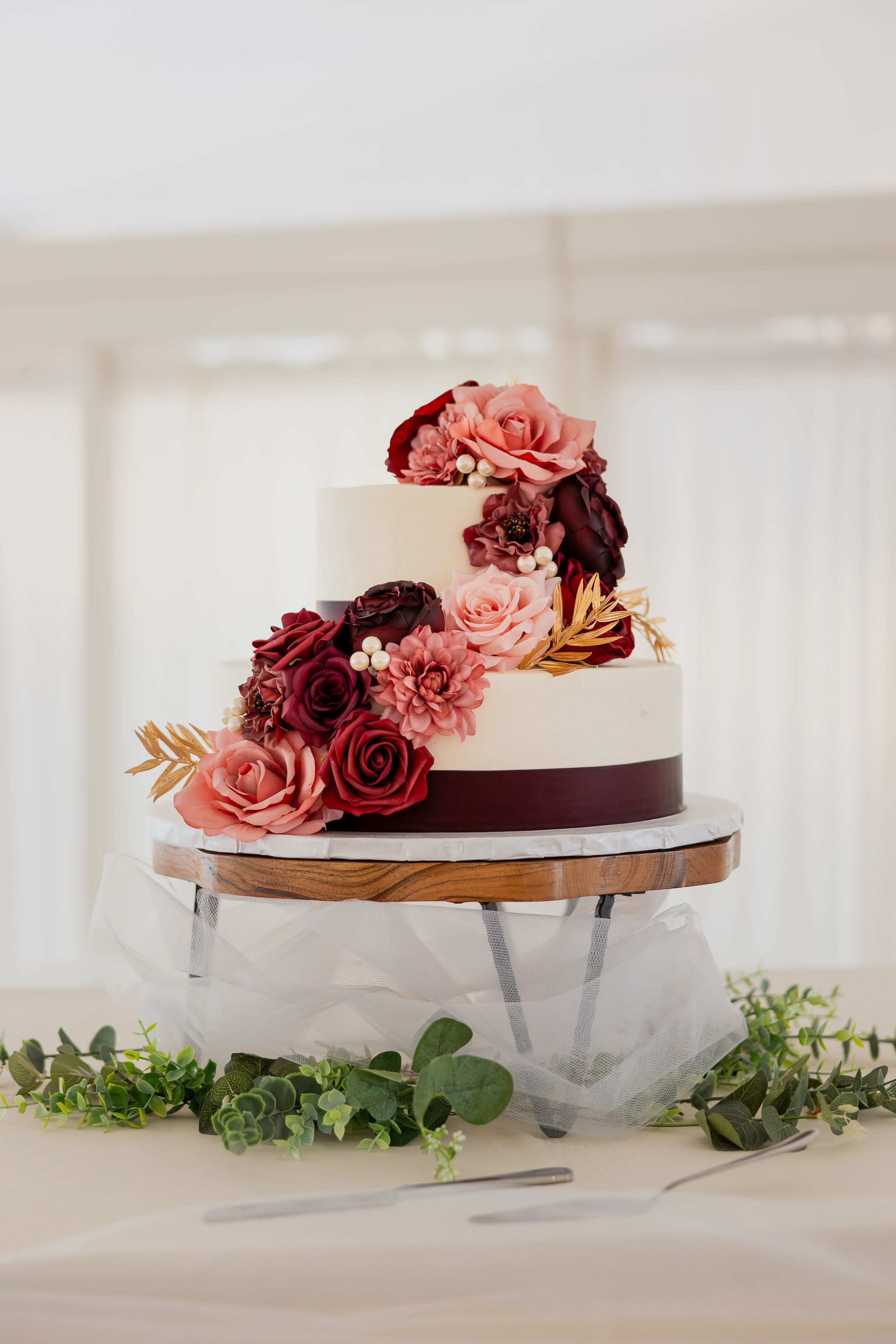 Wedding cake decorated with burgundy and pink roses displayed on wooden stand with eucalyptus greenery.