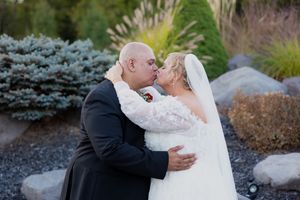 Newlyweds embrace in front of manicured outdoor garden with evergreen trees and stone wall.