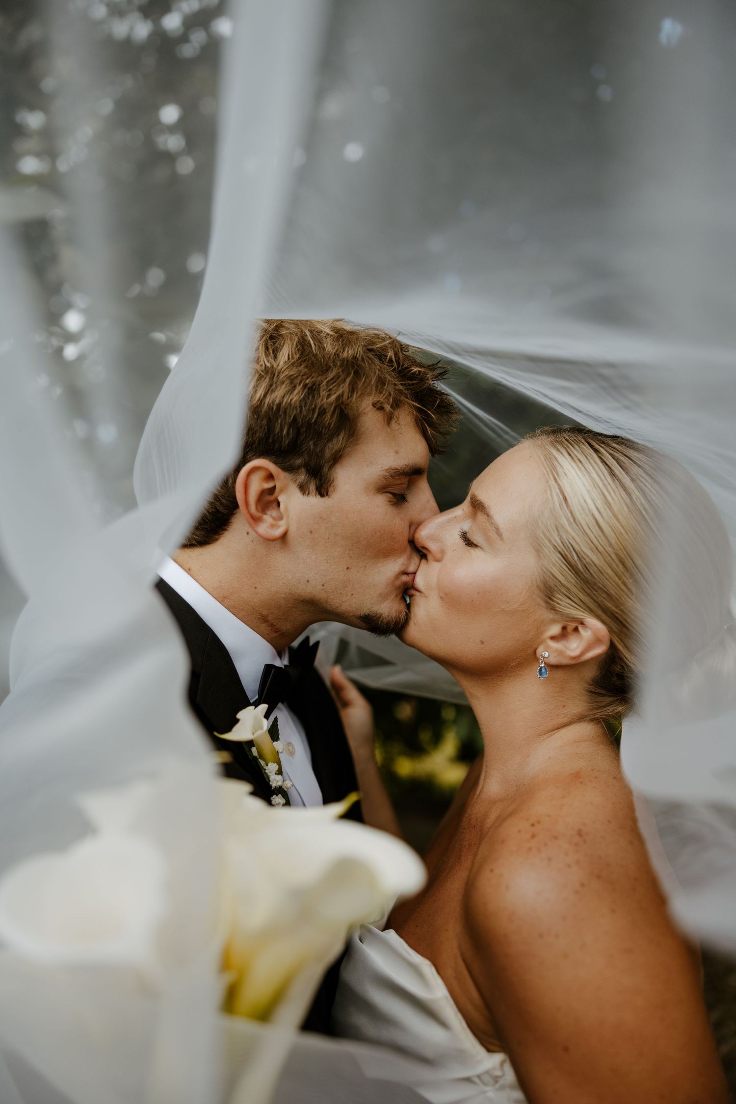Bride and groom sharing a romantic kiss under a flowing white veil with calla lilies in the foreground.