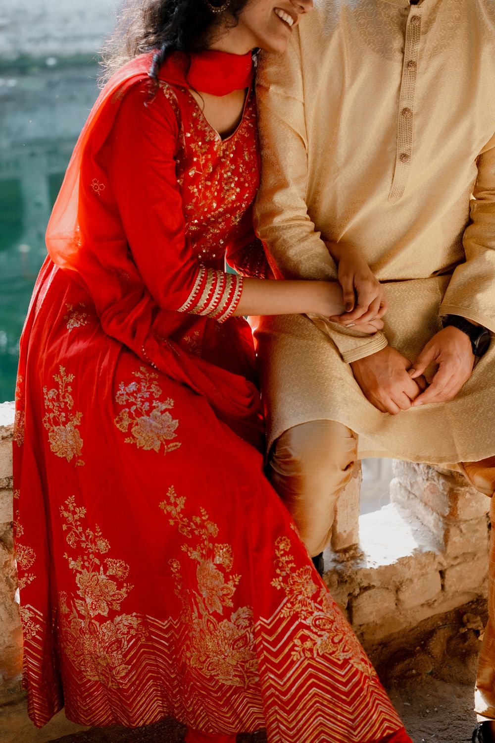 A couple in traditional South Asian attire with a red embroidered lehenga and beige kurta sitting together by the water.