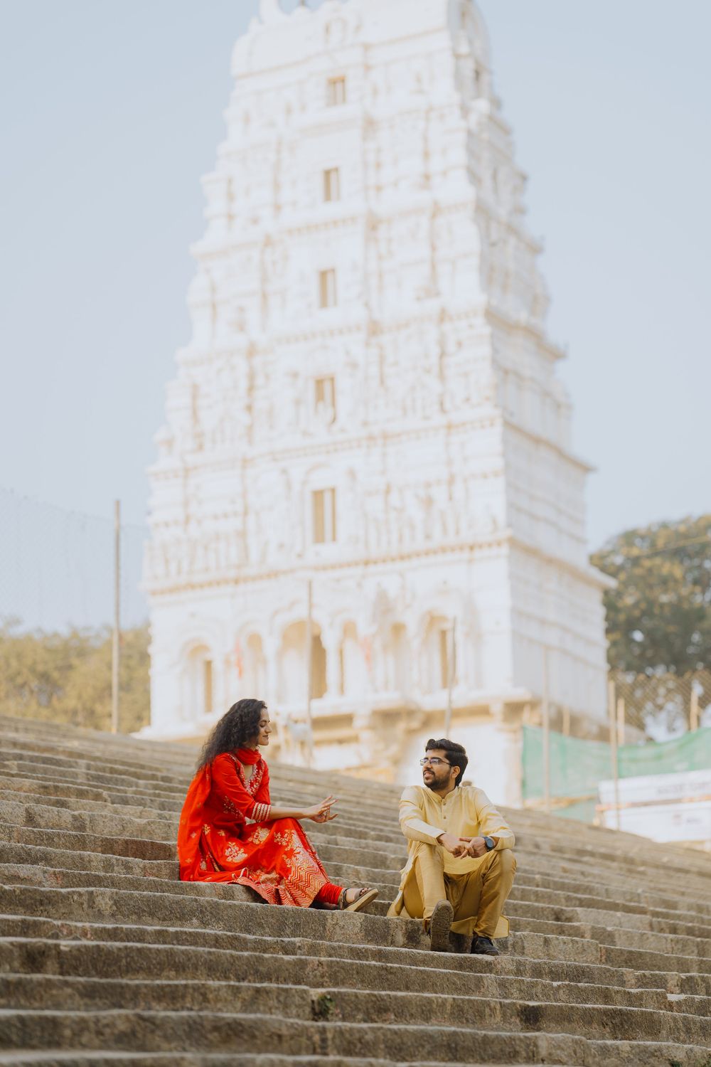 Two people sit and converse on stone steps in front of a white temple tower in traditional South Indian style.