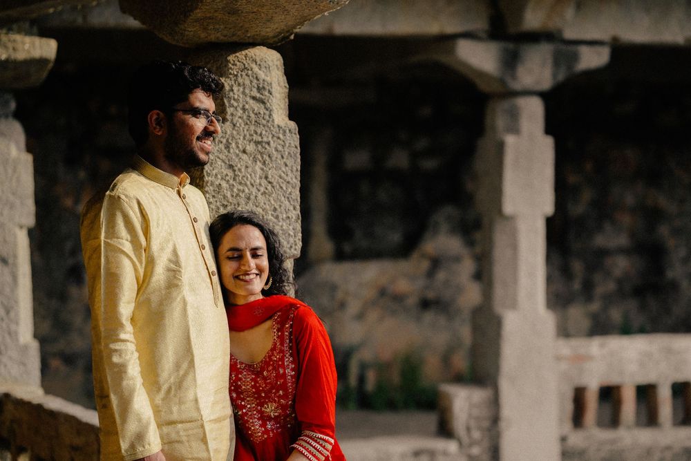 Couple in traditional South Asian attire standing together and smiling under stone pillars.