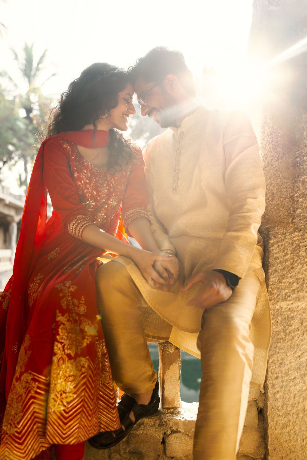 A romantic couple in traditional Indian attire leans against a sunlit wall creating a dreamy silhouette effect.