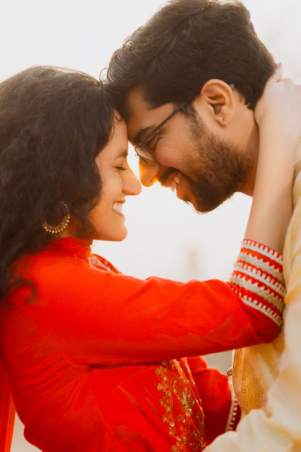 A couple in traditional Indian attire share an intimate moment, with the woman wearing a vibrant red outfit with golden details.