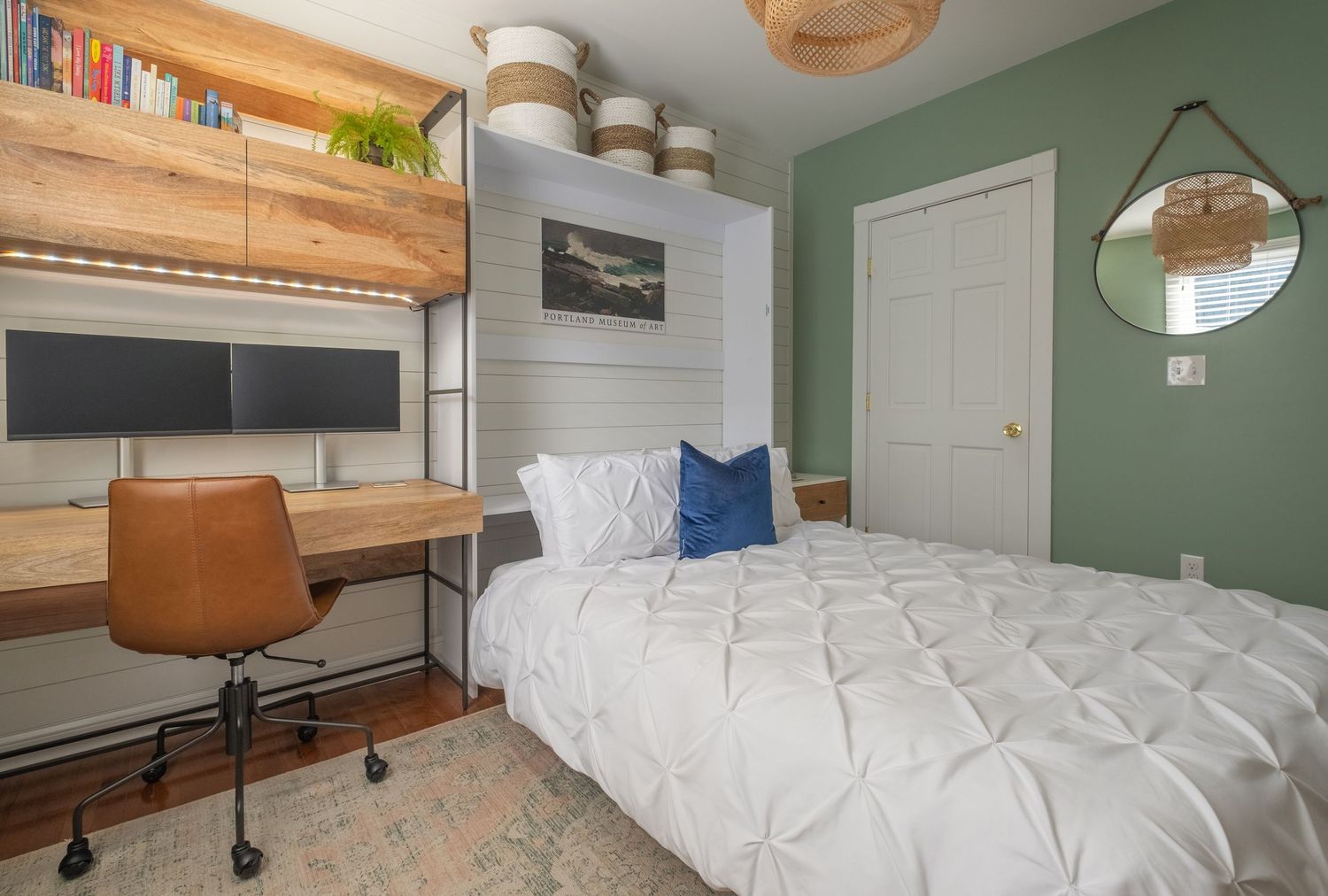 Modern bedroom features a white bed, wooden desk setup, green accent wall, and floating shelves with decor and storage.